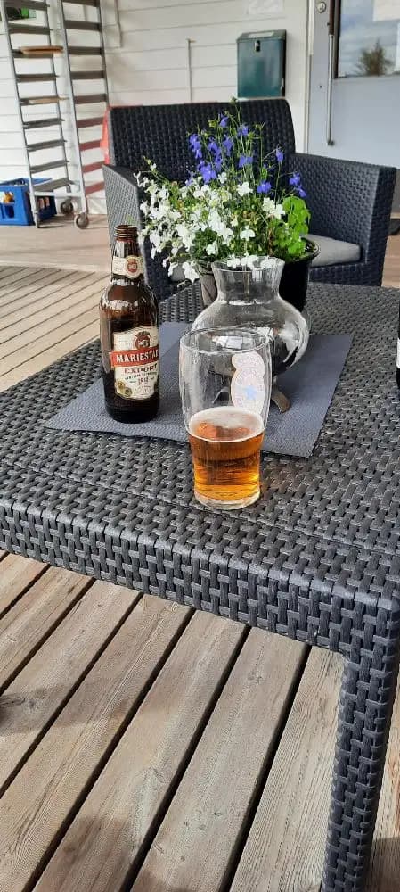 En uteplats vid Borgaruddens havsbad och familjecamping med flätade möbler, inklusive ett bord och stolar. En flaska står på bordet, omgiven av natur och en avkopplande atmosfär.