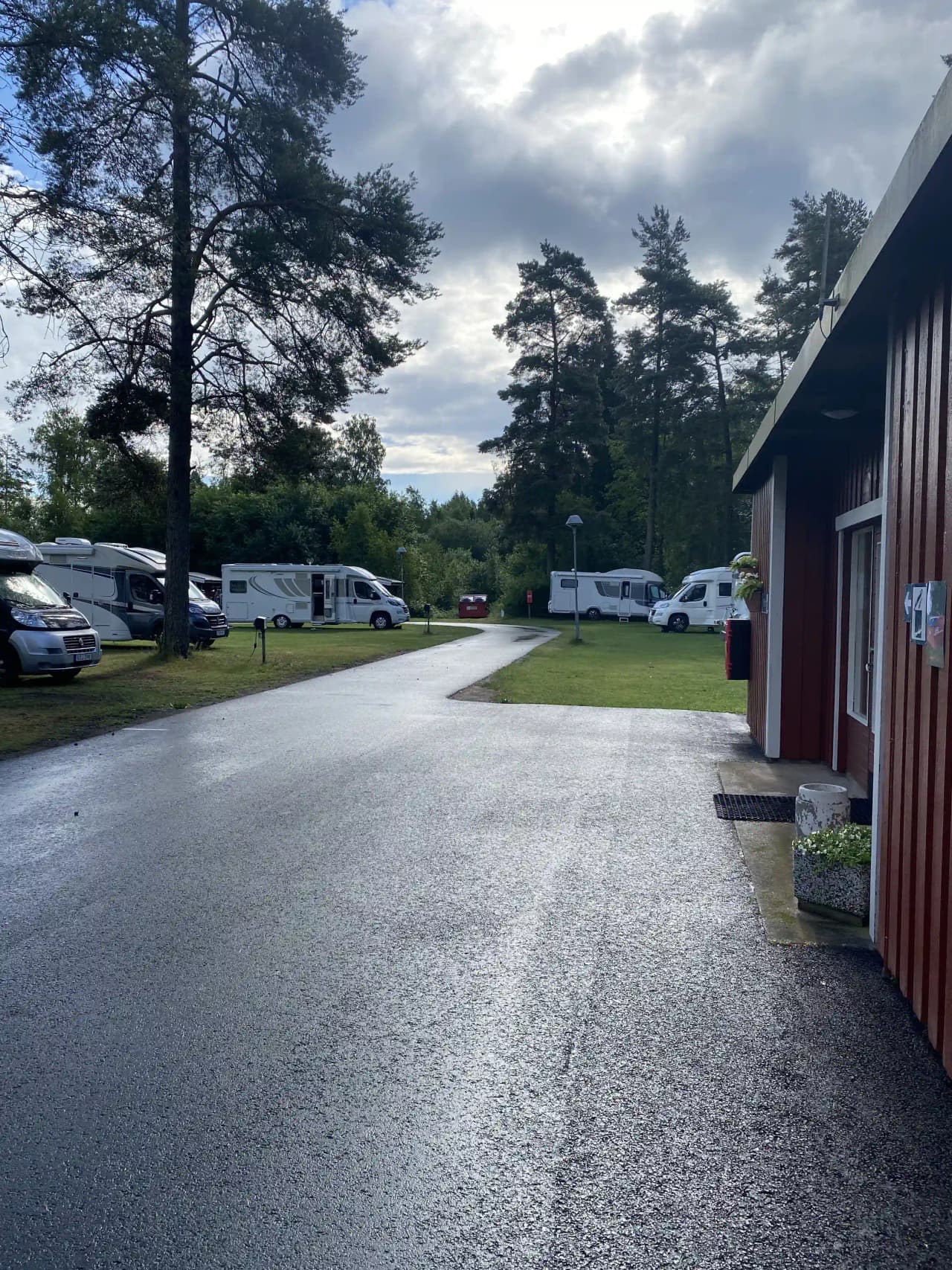 En asfalterad uppfart med parkeringsplatser, där det står ett par bilar, omgiven av natur och belägen vid Hörsjöns camping.