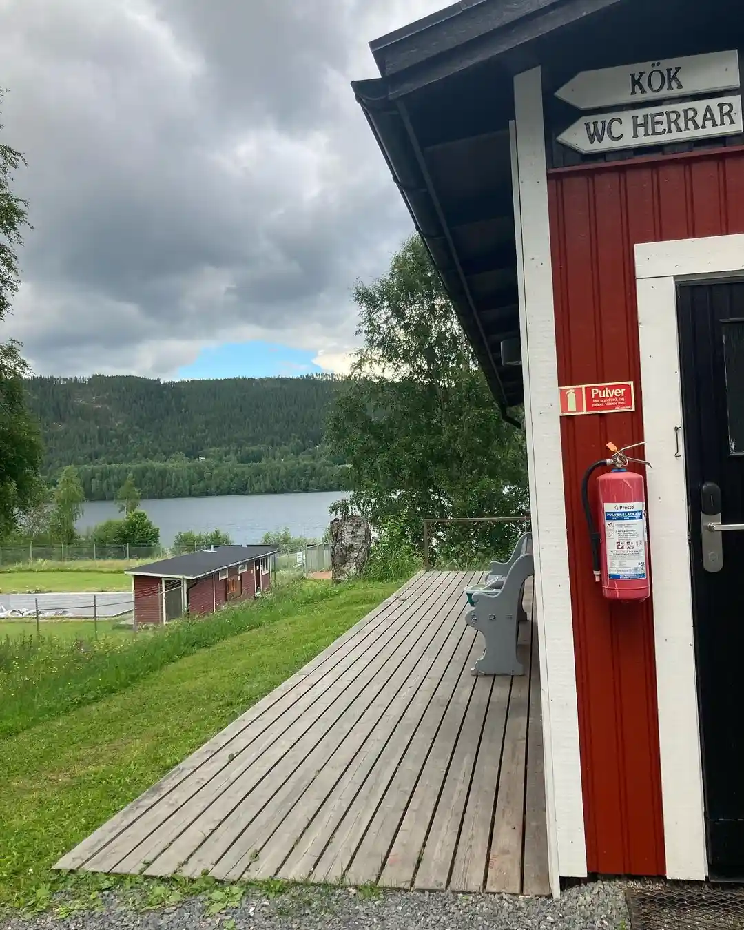 En bild på Hammarstrands camping som visar utemöbler på en gräsmatta. Det finns ett picknickbord och bänkar av mörkt trä, omgivna av grönska.