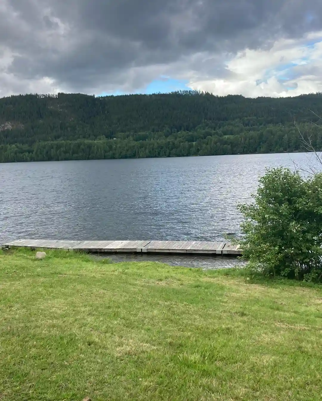 Vy över Hammarstrands camping med en sjö i förgrunden, omgiven av skogsklädda höjder under en delvis molnig himmel.