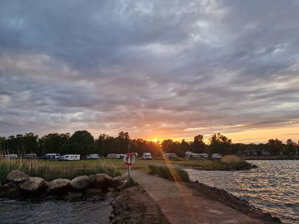 En fridfull vy över en sjö vid skymning med horisonten i bakgrunden, omgiven av en strand och bankar. Solens sista strålar färgar himlen i varma toner vid Björkenäs camping.