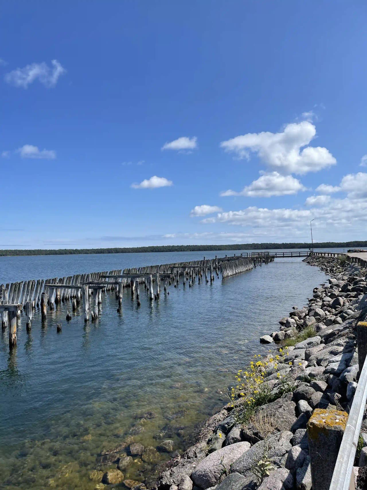 Bild av Böda Hamns Camping med en vidsträckt havsutsikt. Man ser vågor som sköljer in mot strandkanten där det finns en vågbrytare. Horisonten syns i fjärran under en himmel med cumulusmoln.