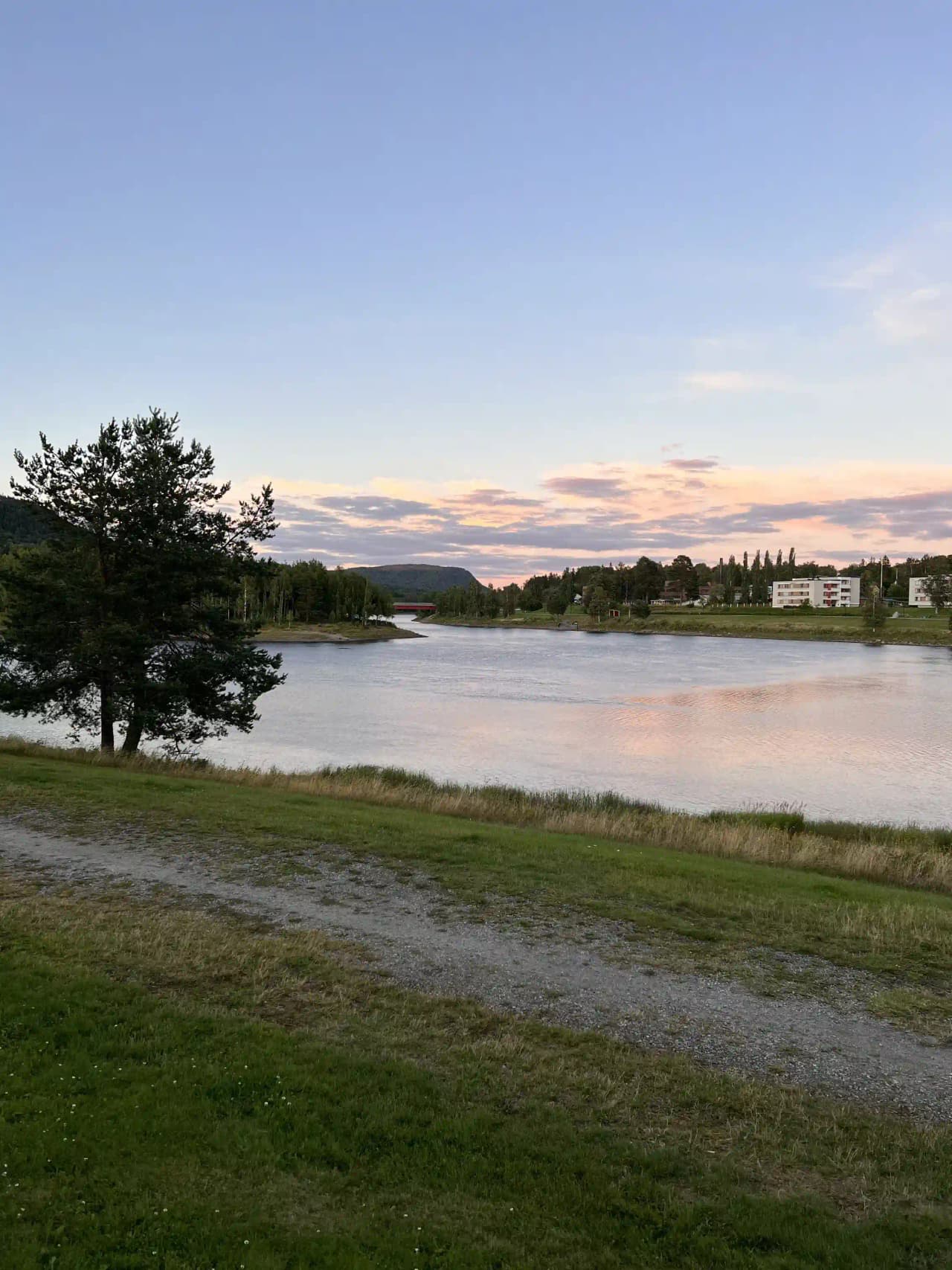En naturskön vy över Sollefteå camping, med en vattenspegel som reflekterar en blå himmel med moln, omgiven av grönska och naturlandskap i horisonten.