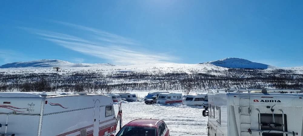 En snöig vintermiljö vid Fjällnäs camping med bergskedja i bakgrunden. Flera husbilar och vanar står parkerade, täckta av snö och is.