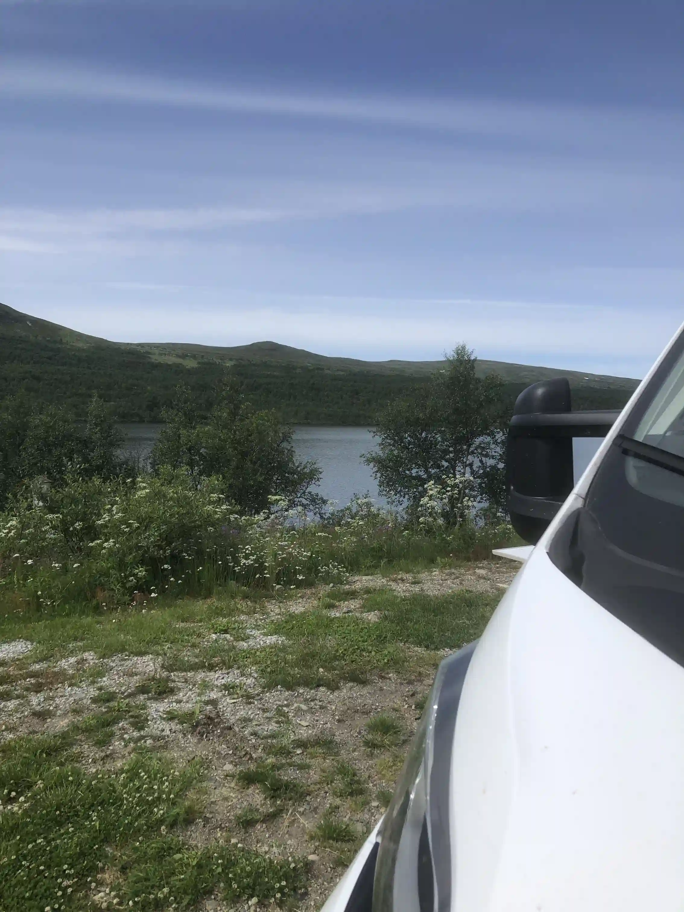 En bil står parkerad vid fjällnäs camping med utsikt över vidsträckta fjäll under en himmel med spridda moln. Bilens sidobackspegel syns i förgrunden.