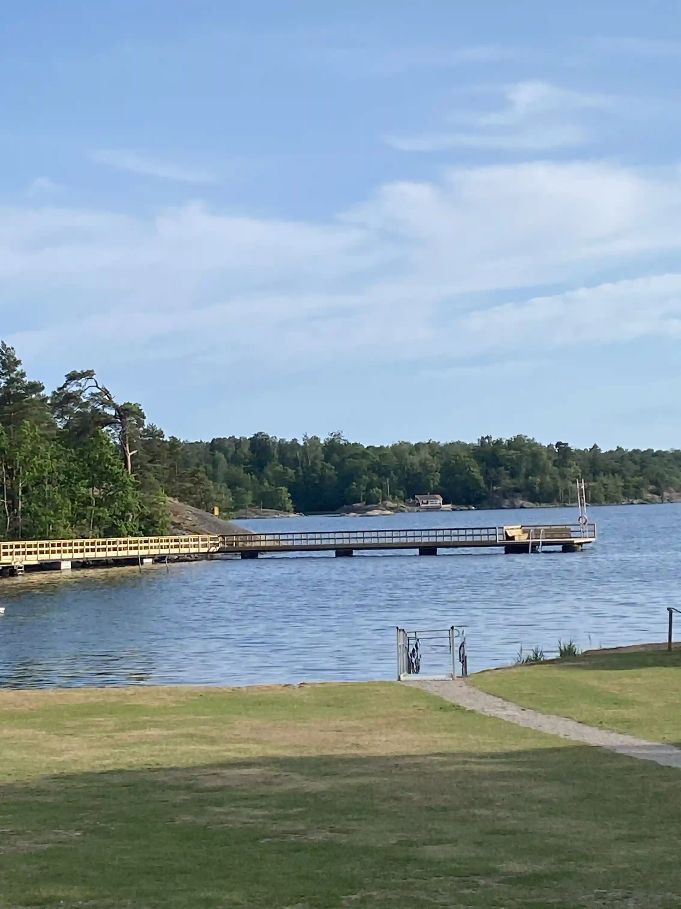 En vy över Järnaviks camping med vatten i förgrunden, där en brygga sträcker sig ut i vattnet. Bakom bryggan syns en grönskande strandbank vid sjöns eller kanalens kant, omgiven av natursköna kustlandskap.
