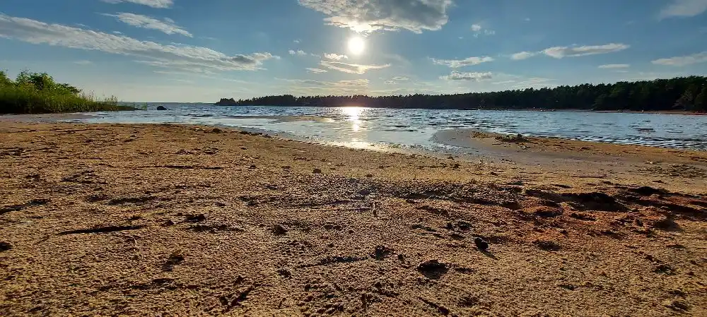 En naturskön vy över Bomstadbaden Camping visar en lugn vattenyta vid horisonten, omgiven av moln och en naturlig strandlinje.