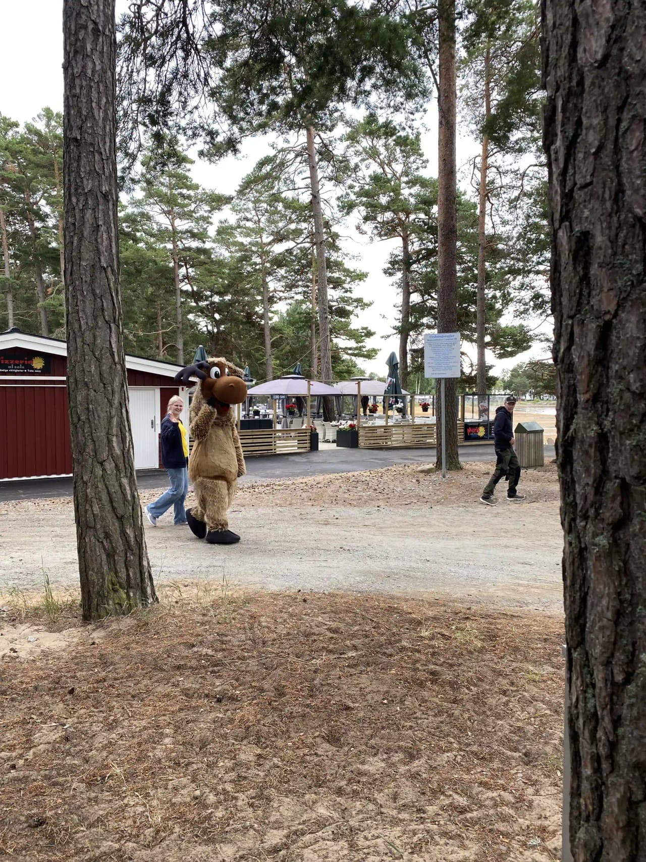 En maskot poserar vid ingången till Bomstadbaden camping, omgiven av gröna träd och en upplyftande atmosfär av rekreation och utomhusaktiviteter.