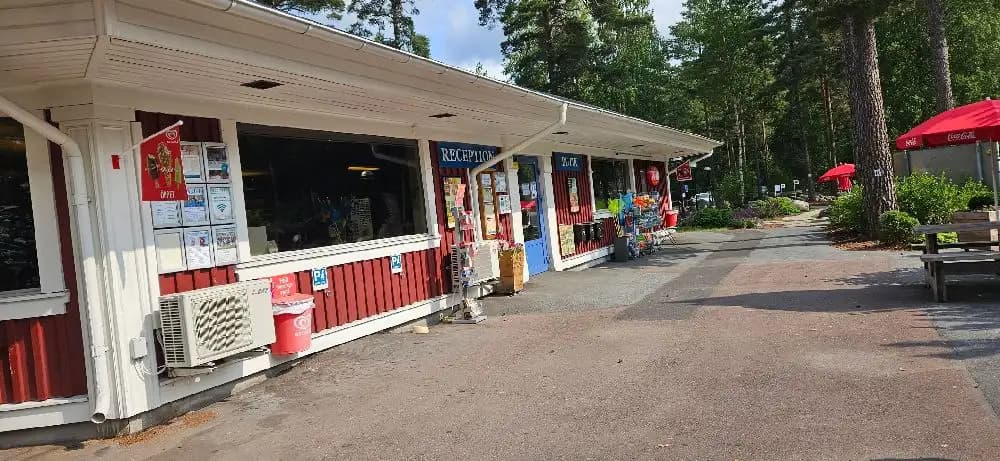 En skuggig gångväg med bänkar och blomkrukor vid Bomstadbaden Camping, intill en restaurang och ett vilaområde.