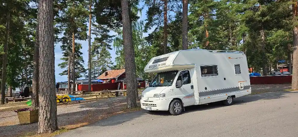 En husbil parkerad på Bomstadbaden camping, omgiven av andra campingfordon och träd. I förgrunden syns bilens hjul och registreringsskylt.