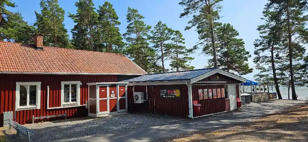 En timmerstuga i skuggan av träd med ett picknickbord och utemöbler på gräsmattan vid Bomstadbaden camping.