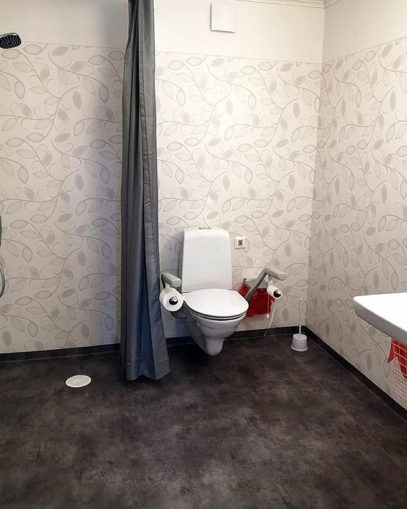 Bild av ett badrum med ljusgrå kaklade väggar och golv. Rummet är utrustat med en vit toalettstol och ett enkelt, stilrent handfat.