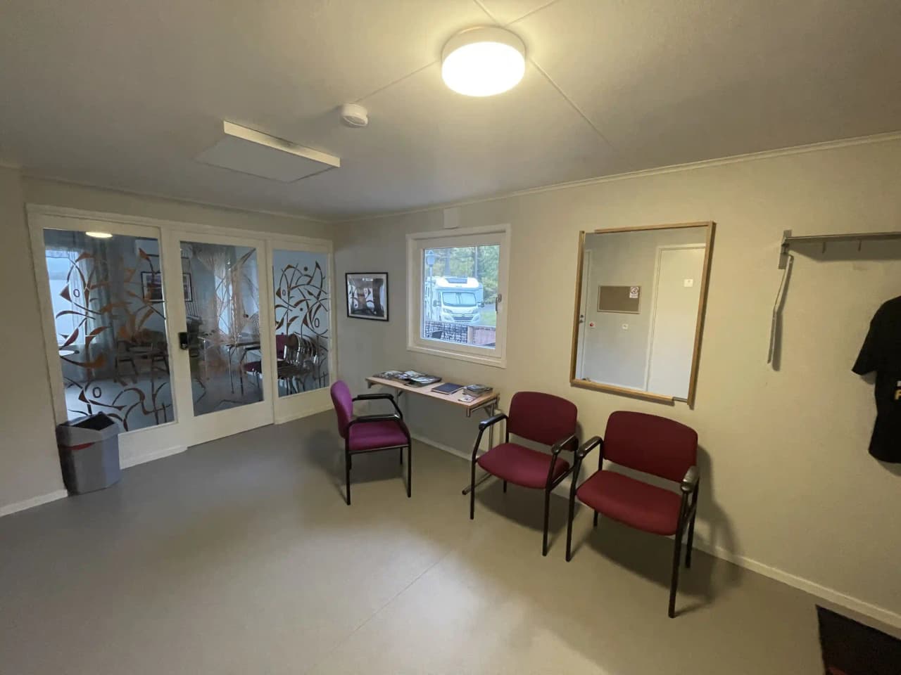 En ljus och modern inomhusmiljö med golv i trä och tak med inbyggda lampor. Rummet är möblerat med stolar och har en inramad bild på väggen. Det ser ut som en vänthall med fokus på trivsam och rymlig interiördesign.