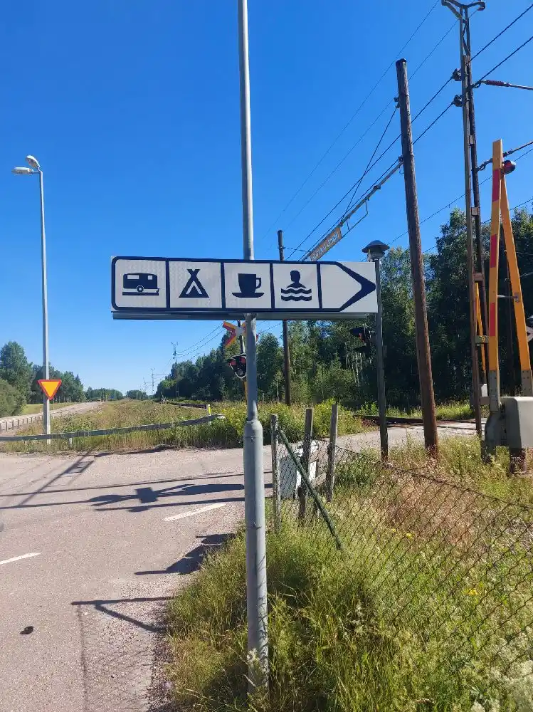 En bild av Horndals bad & camping med en tydlig kraftledning och en skylt vid sidan av vägen. Området inkluderar infrastruktur som trafik- och gatunamnsskyltar i förgrunden, medan bad- och campingområdet ligger i bakgrunden.