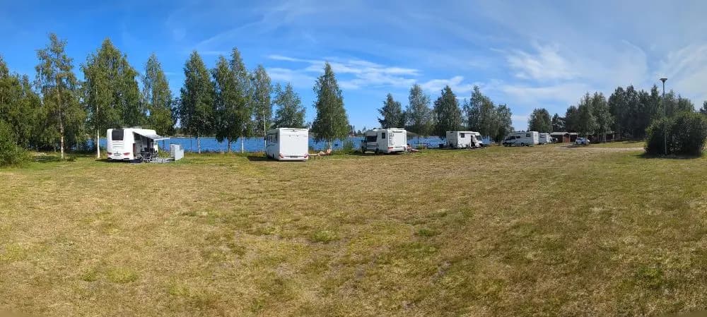 En vidsträckt gräsplan på Sikeå Havscamping med flera husvagnar och husbilar parkerade. En semi-trailer lastbil syns i periferin, omgiven av prärie- och ängslandskap.