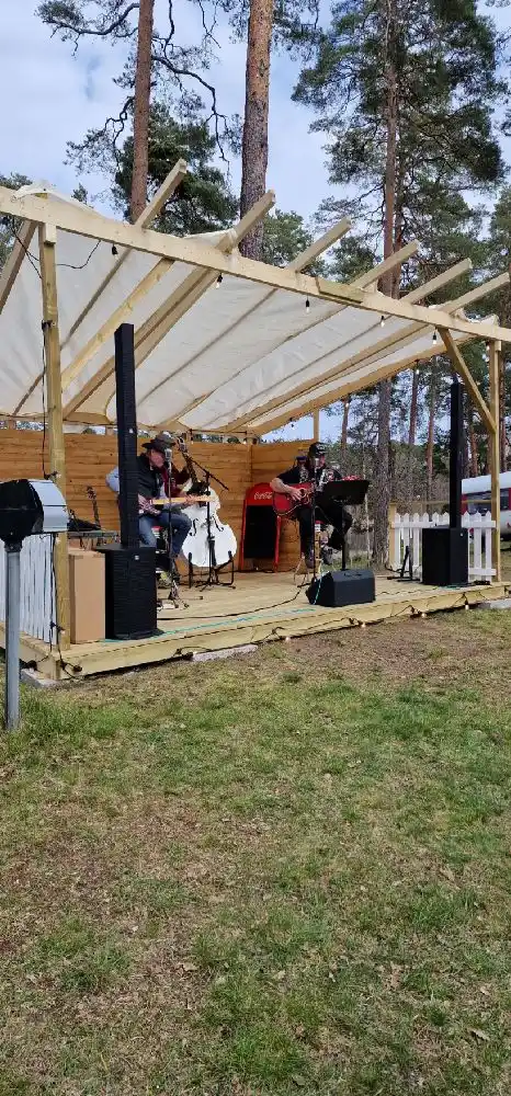En campingplats omgiven av tät skog visar en utomhusscen med paviljongliknande struktur under trädkronorna, perfekt för framträdanden och samlingar.
