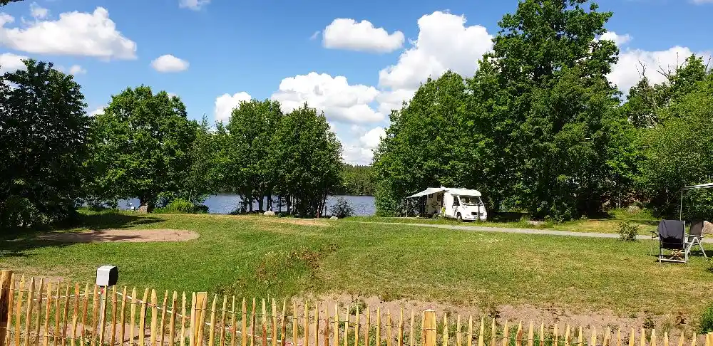 Bild av Långsjön Stugor & Camping: En grönskande campingplats med träd, gräs och en damm. Det finns en picknickbänk placerad på en gräsmatta, omgivet av ett naturskönt landskap med ängar och trädgårdsväxter.