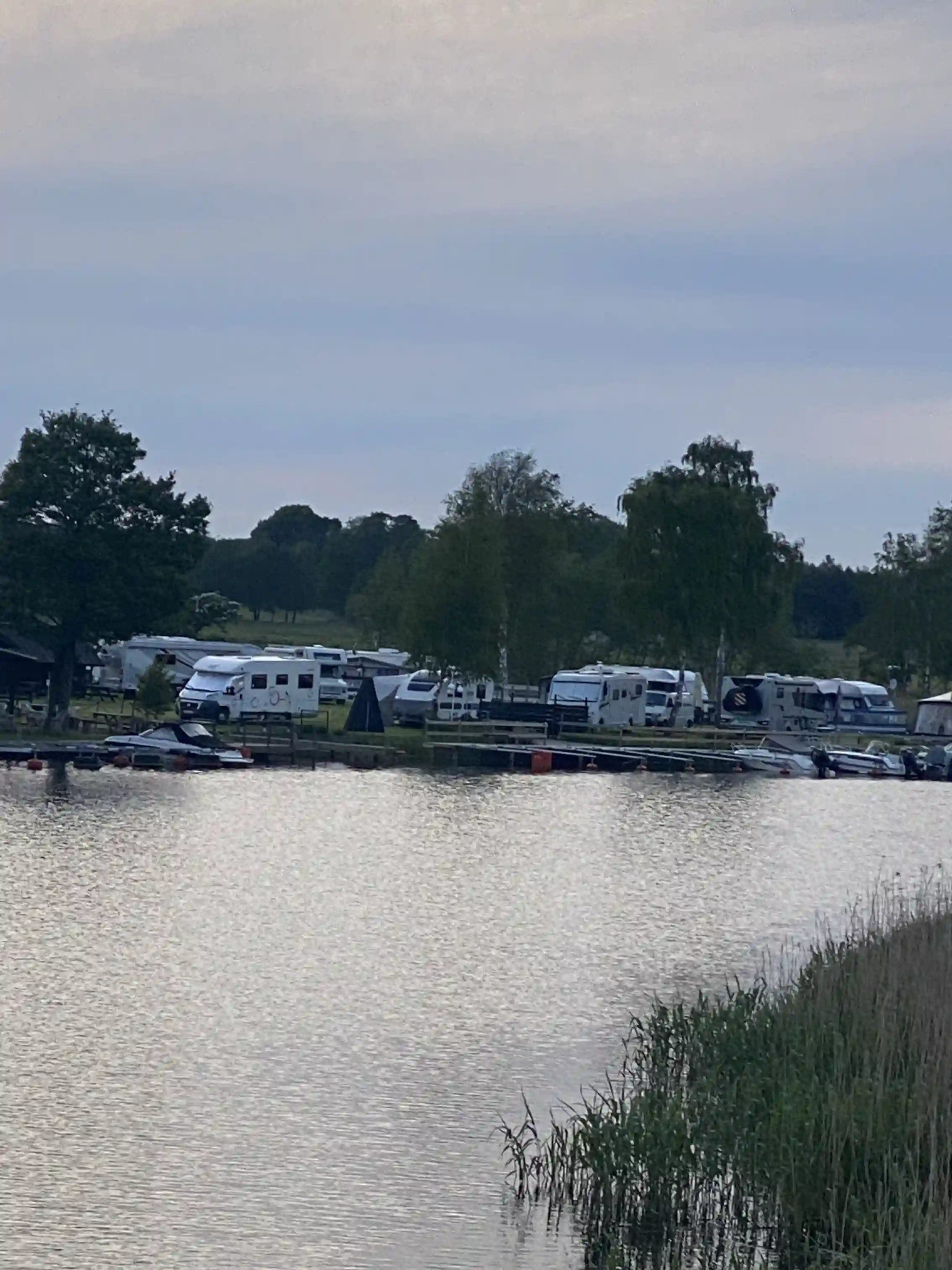 En bild av en stilla vattenväg vid Martins Rökeri och Camping, med speglingar från den omgivande naturen. Flera båtar ligger förtöjda längs strandbanken.