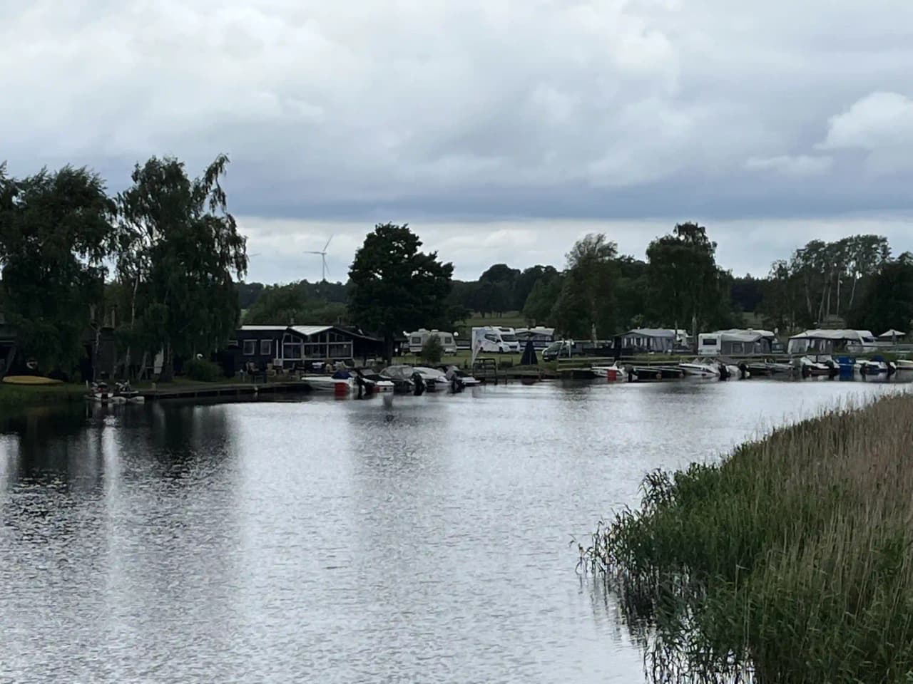 En campingplats vid en sjö, där en rök från Martins rökeri syns i bakgrunden. Flera båtar ligger förtöjda vid strandkanten under en molnig himmel.