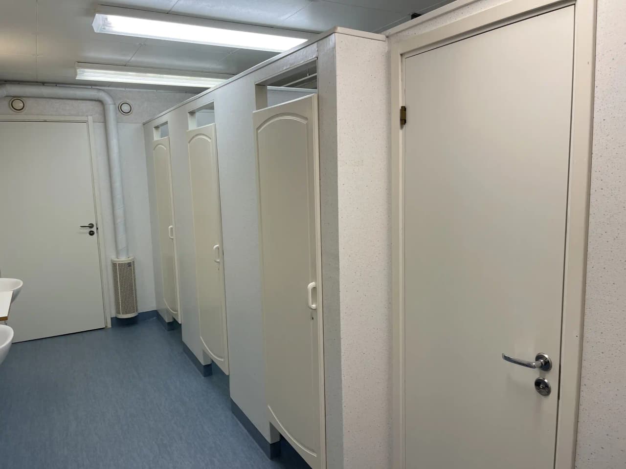 En enkel toalett med klinkergolv och vitmålade väggar, utrustad med en dörr, handfat och en toalettstol. Toaletten finns på Martins Rökeri och Camping, ett besöksmål för campingentusiaster.