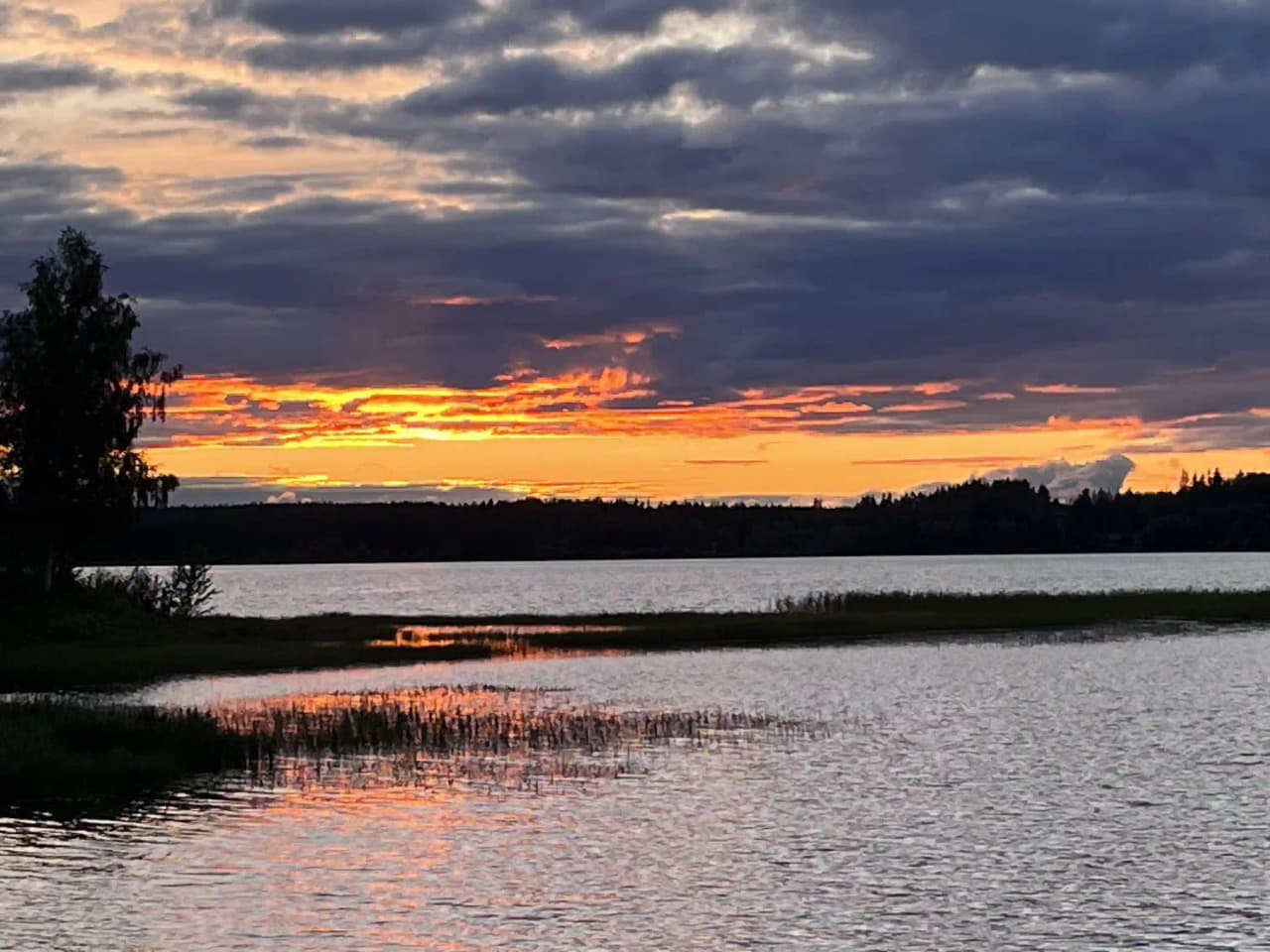 En rofylld kvällsutsikt vid Särna camping med en spegelblank sjö i förgrunden, omgiven av en klar blå himmel och svagt upplysta moln vid horisonten i skymningsljus.