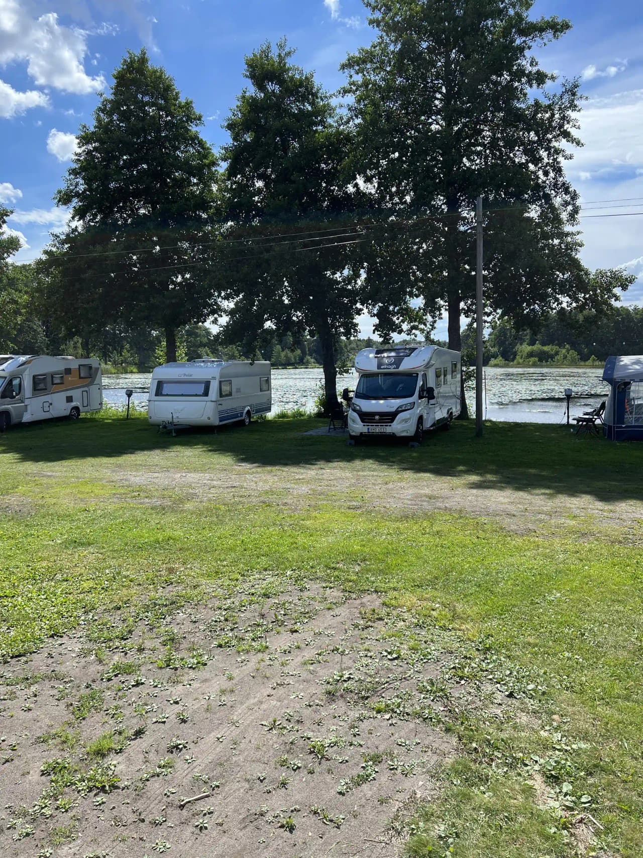 En campingplats med husbilar och husvagnar parkerade på en gräsbevuxen yta, omgiven av träd och med en asfalterad parkeringsplats i närheten.