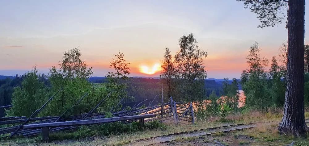 Solnedgång över Näsåkers camping & stugby, där solen sänker sig bakom en naturlig landsbygd med öppna ytor och skogskanter, vilket skapar ett lugnande ljus över platsen.
