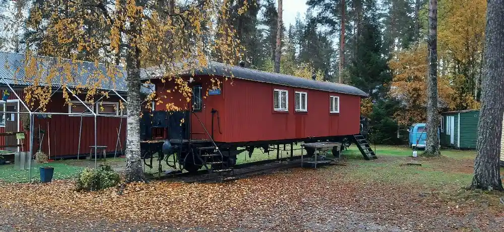 En träbeklädd järnvägsvagn står på en gräsbevuxen gård vid Näsåkers camping & stugby, omgiven av några gamla trähjul och järnskrot. I bakgrunden syns skog och kullar under en klarblå himmel.