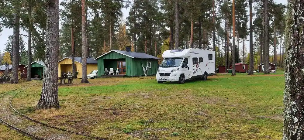 En grönskande campingplats med träd i bakgrunden. I förgrunden syns stugor på en gräsmatta, en husvagn och en uppfart. Perfekt för natur- och friluftsentusiaster.