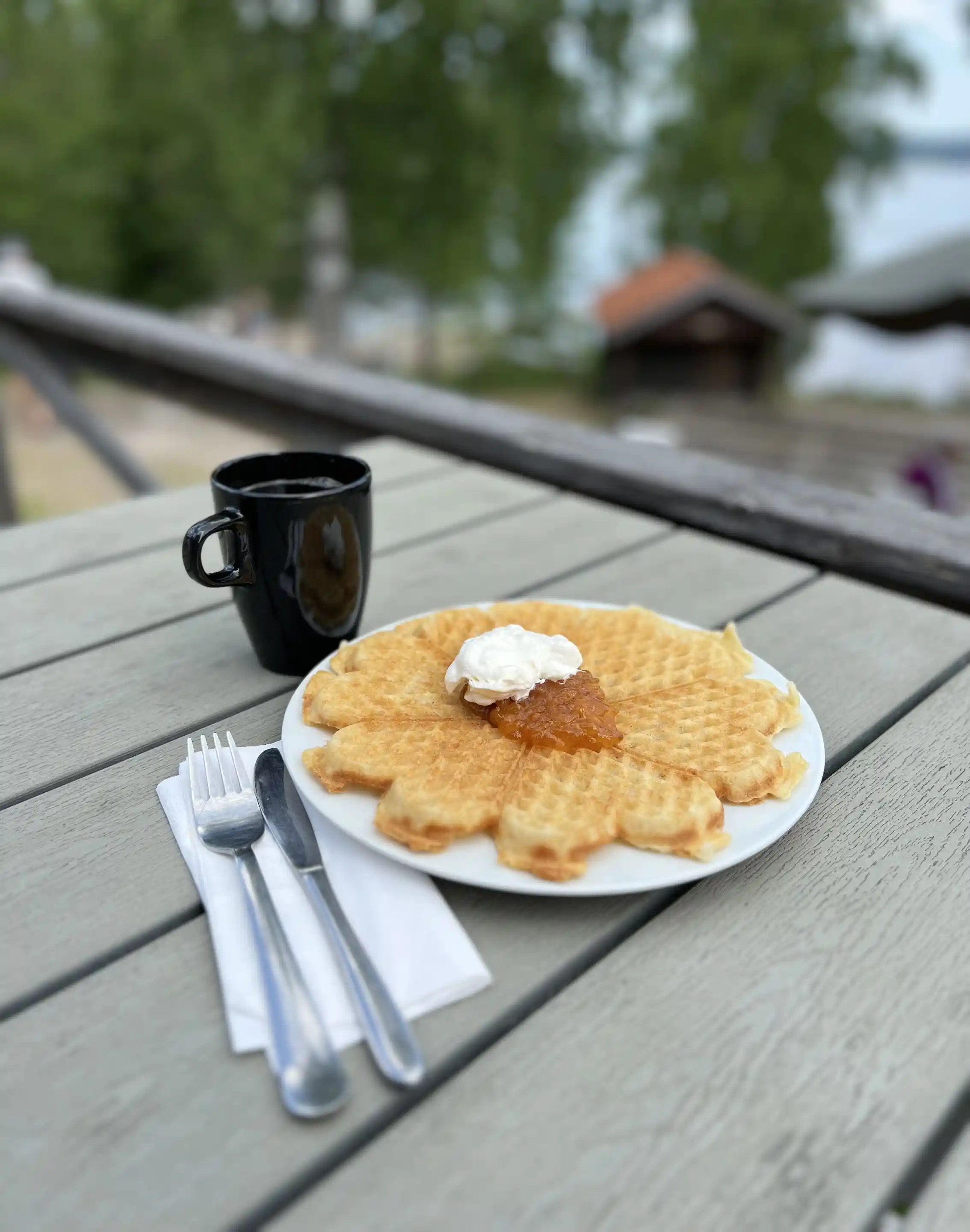 En tallrik med nygräddade våfflor toppade med grädde och bär, omgiven av bestick och glas på ett träbord.