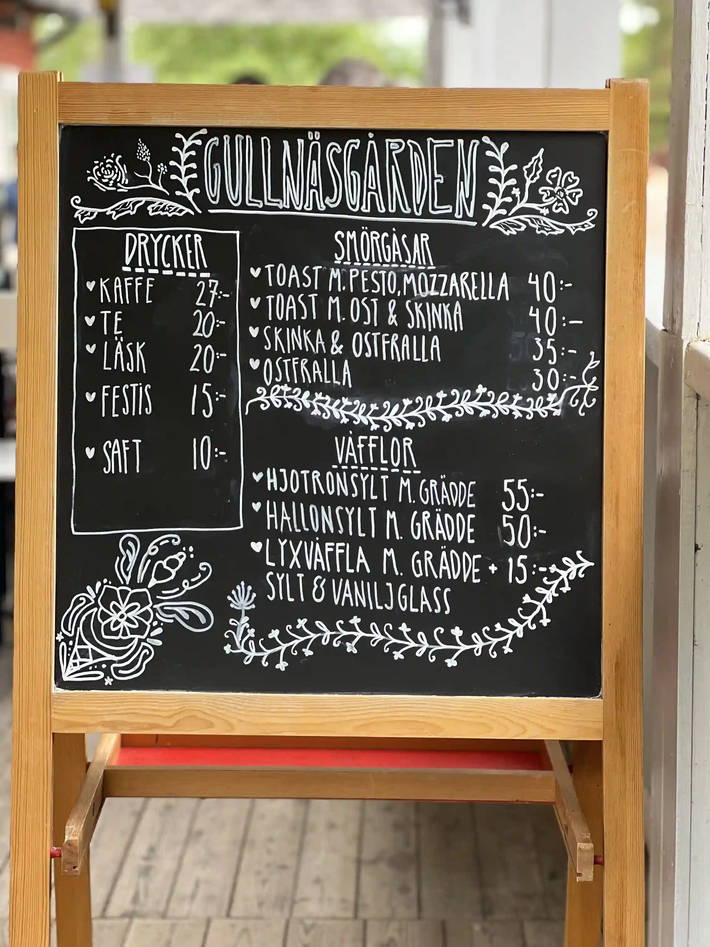 En svart tavla med handskriven meny i krita står bredvid några trästolar och ett bord, vilket ger en inbjudande och hemtrevlig atmosfär på Gullnäsgården.