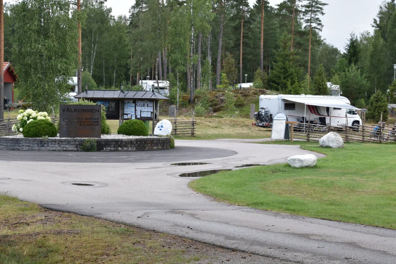 En grönskande trädgård med uppfart omgiven av växter och frodig landskapsplanering. En husbil är parkerad i närheten, med en stolpe synlig på avstånd.