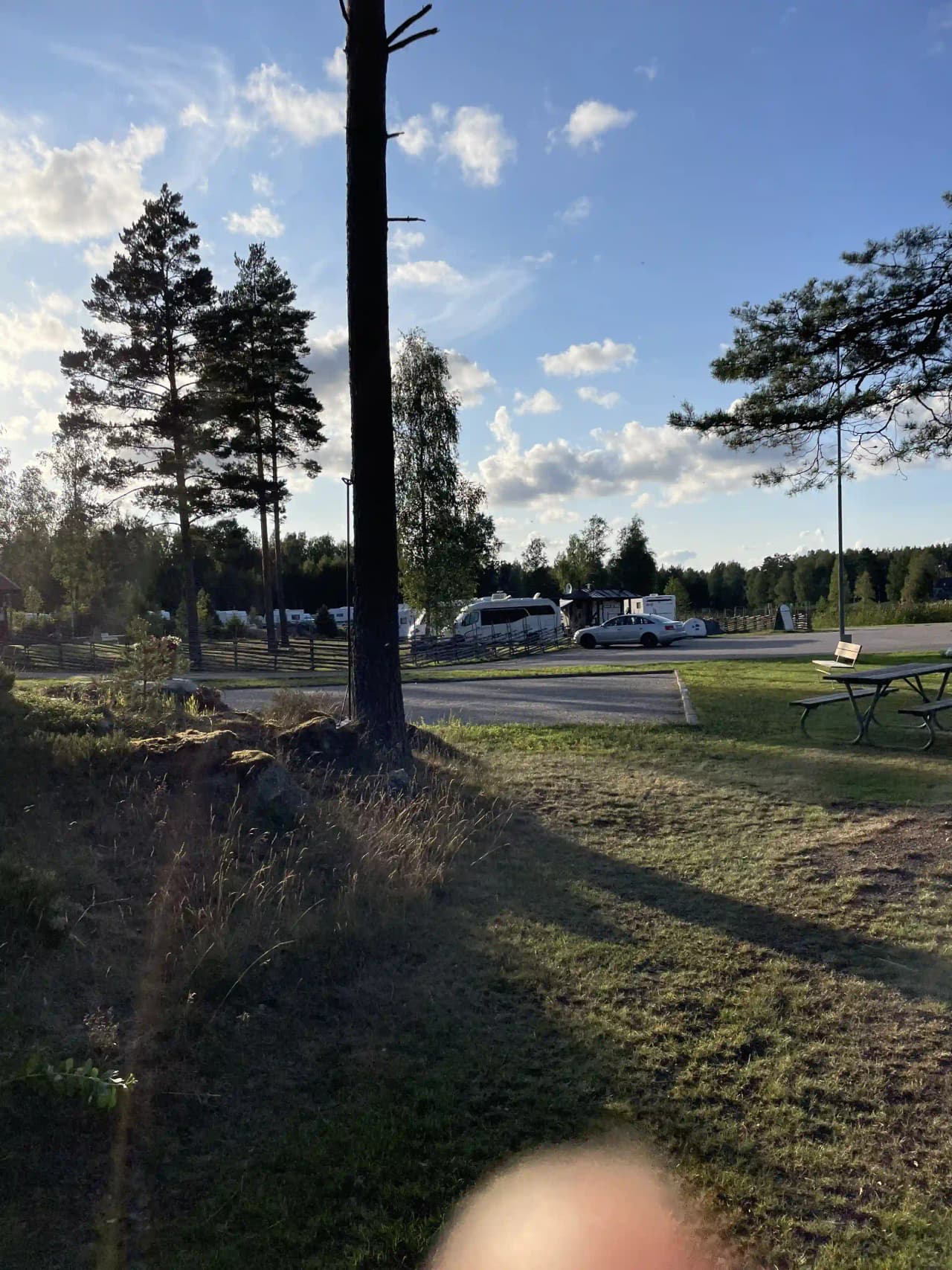 En vidsträckt vildmarkscamping med öppna slätter, omgivna av barrträd under en himmel med cumulusskyar. En enskild stolpe syns mot himlen.