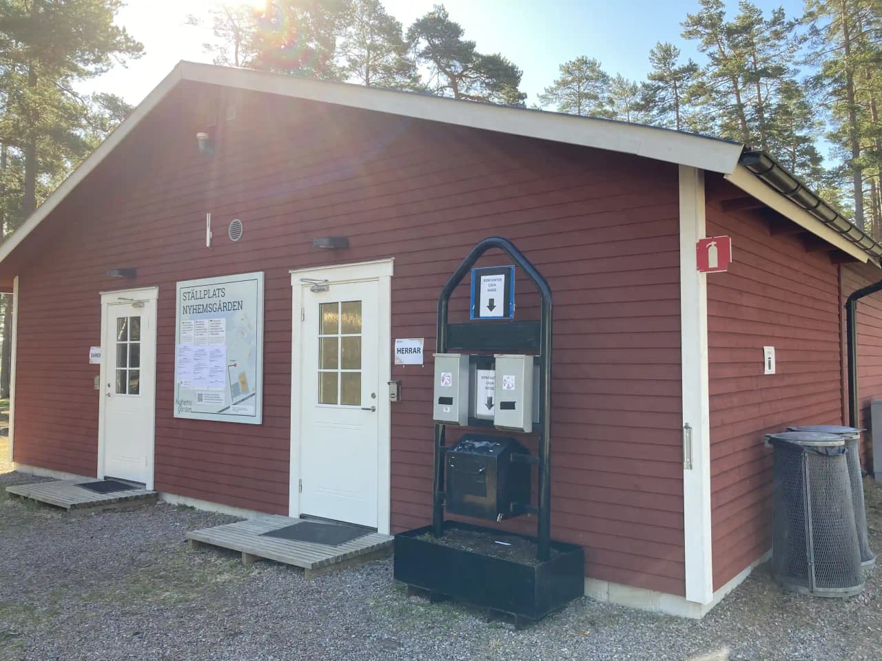 En trästuga med en träfasad och en dörr, omgiven av en gårdsplan och en uppfart. Byggnaden har en stugliknande design och är placerad i en lantlig miljö.