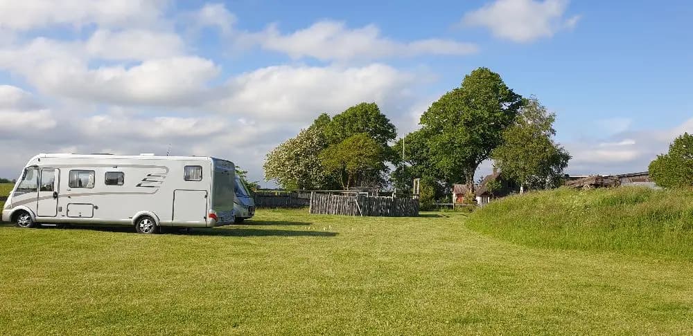 En husbil och en husvagn står parkerade på en grön gräsmatta på en ställplats i ett parkliknande område.