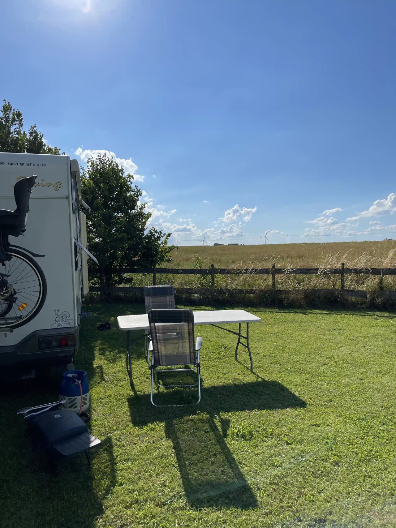 En grön gräsmatta med flera husbilar och husvagnar uppställda. Utomhusmöbler som bord och stolar är placerade i skuggan bredvid fordonen. miljön ger en trivsam campingkänsla.
