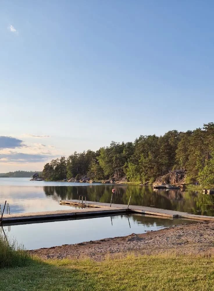 En brygga sträcker sig ut över en stilla sjö vid Tyrislöt Camping, omgiven av lummig grönska och reflekterande vatten.