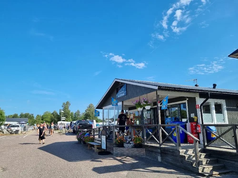 En livlig campingplats vid Sandbybadet med tält och cyklar parkerade under skuggande träd. Människor i grupp njuter av fritidsaktiviteter och samvaro i det offentliga utrymmet.