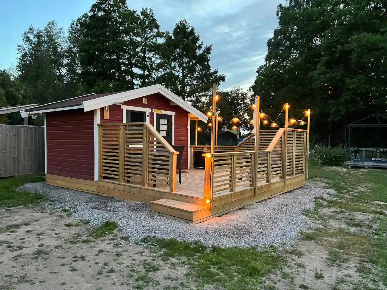 En trästuga med altan omgiven av en grönskande natur, som finns vid Mälarbadens camping. Trappor leder upp till huvudentrén, och stugan är byggd av träplankor med en varm träbets.