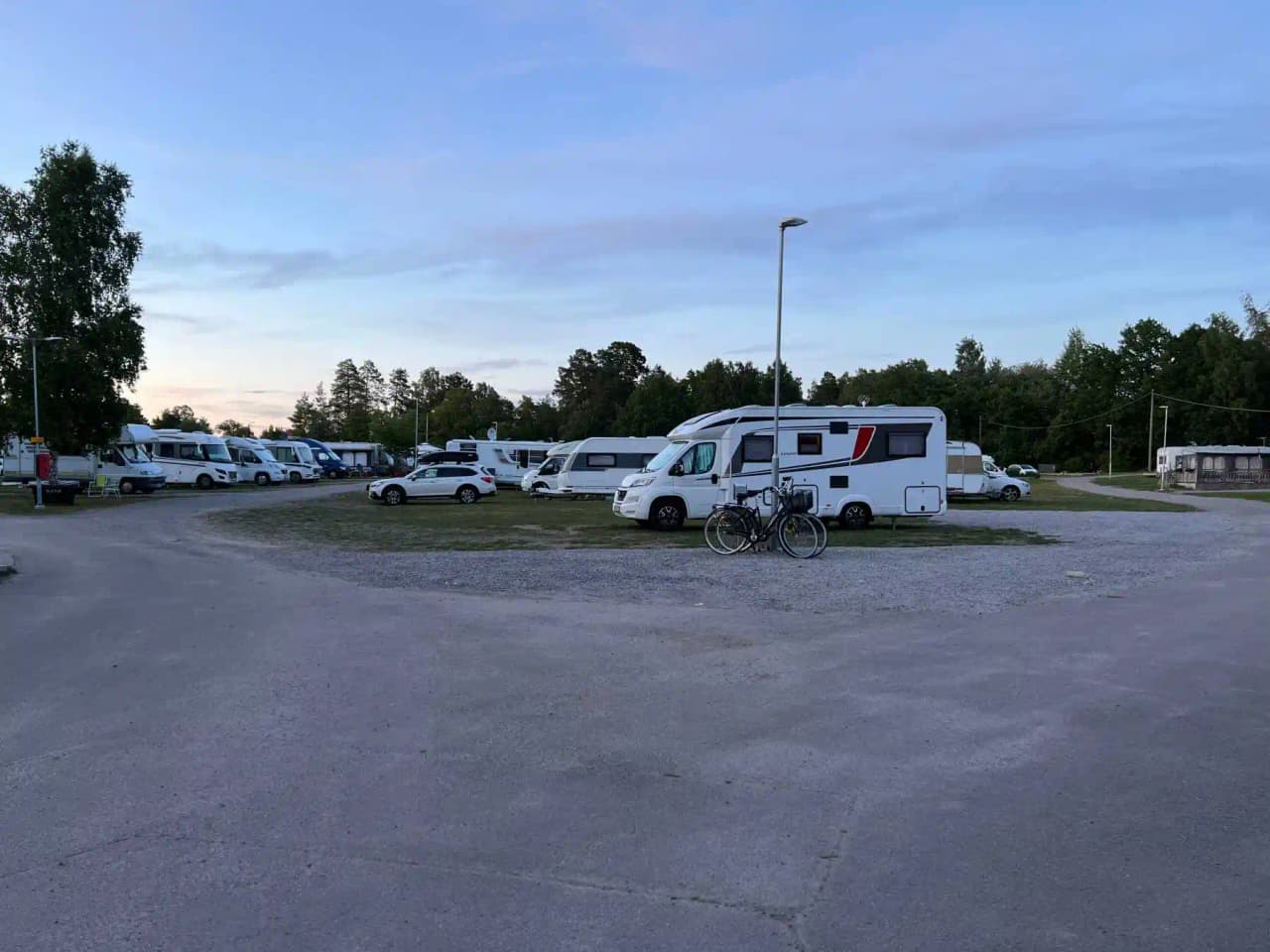 En stor grusparkering vid Mälarbadens camping med flera husbilar, personbilar, skåpbilar och en husvagn. I bakgrunden syns skog och en klar himmel.