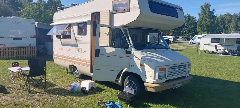 En husbil parkerad vid Mälarbadens camping, med klarblå himmel i bakgrunden och grönska runtomkring.