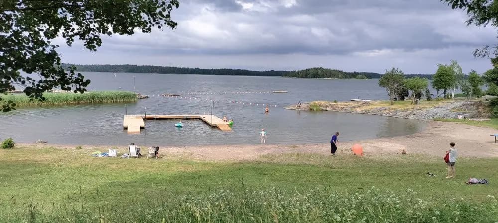 En naturskön vy över Hargshamns camping, med en glittrande sjö i förgrunden omgiven av grönskande skogslandskap. Perfekt för rekreation och avkoppling vid kusten.