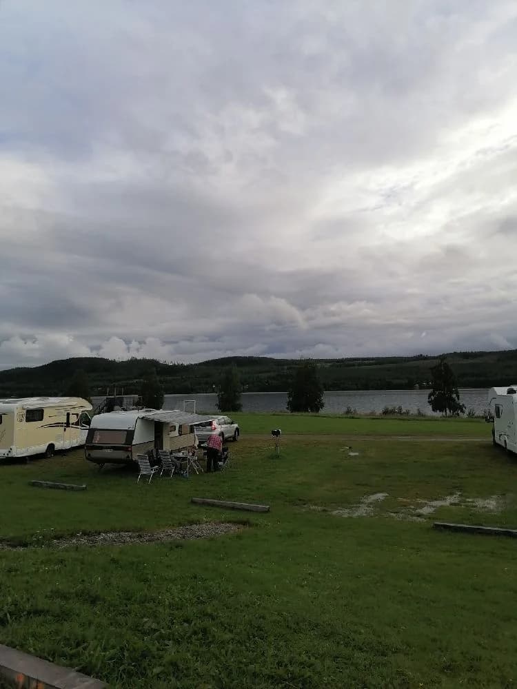 En campingplats vid en sjö med husvagnar och en husbil, ett picknickbord står vid kanten av vattnet under en himmel med fluffiga cumulusmoln.