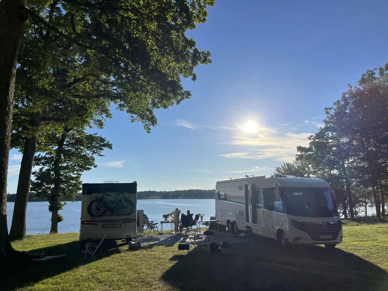 En husvagn står parkerad på en grön gräsmatta i en campingmiljö, omgiven av träd och öppna ytor. I bakgrunden syns ett stort tält och en halvlång lastbil, vilket ger en känsla av friluftsliv och naturlig skönhet i området kring Hägern Bed & Nature.