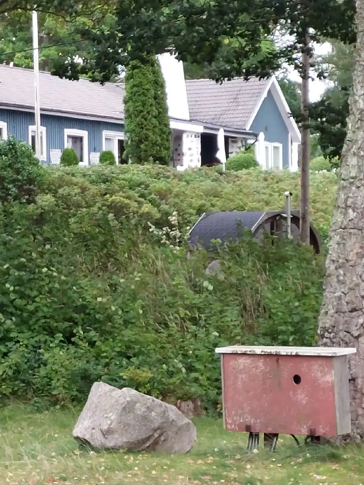 Bilden visar en grön och frodig trädgård vid Hägern Bed & Nature. Omgiven av buskar och marktäckande växter står en pittoresk stuga. Trädgården har en välskött gräsmatta och olika trädgårdsanläggningar med barrväxter, vilket ger en lugn och naturnära atmosfär.