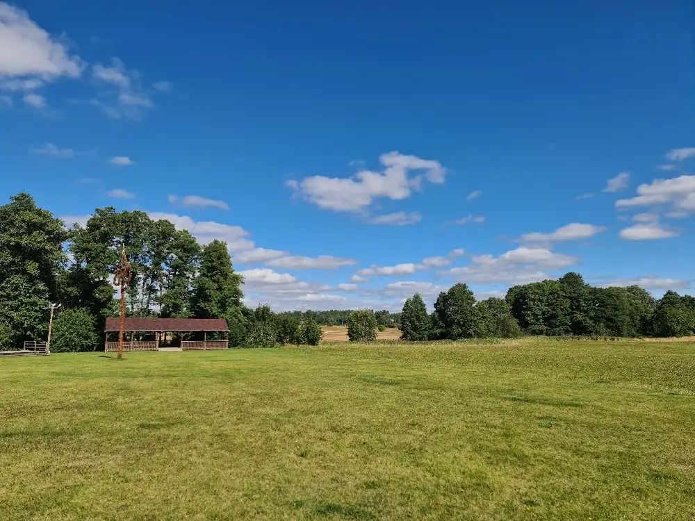 En grönskande hembygdsgård med gröna träd och ett öppet gräsfält under en blå himmel med moln.