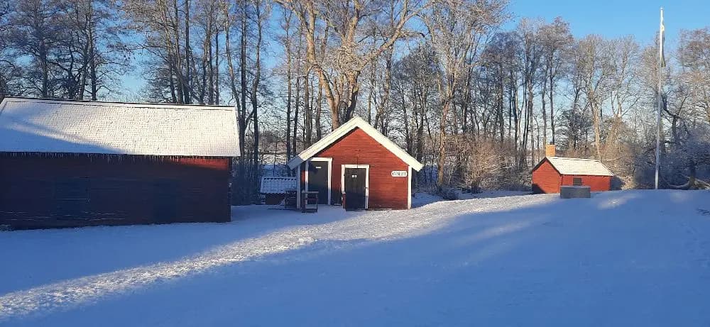 En vit vinterlandskap med snötyngda träd omger kvarngården hembygdsgård. Det lilla trähuset har ett snötäckt tak, vilket ger en charmig och fridfull stämning.