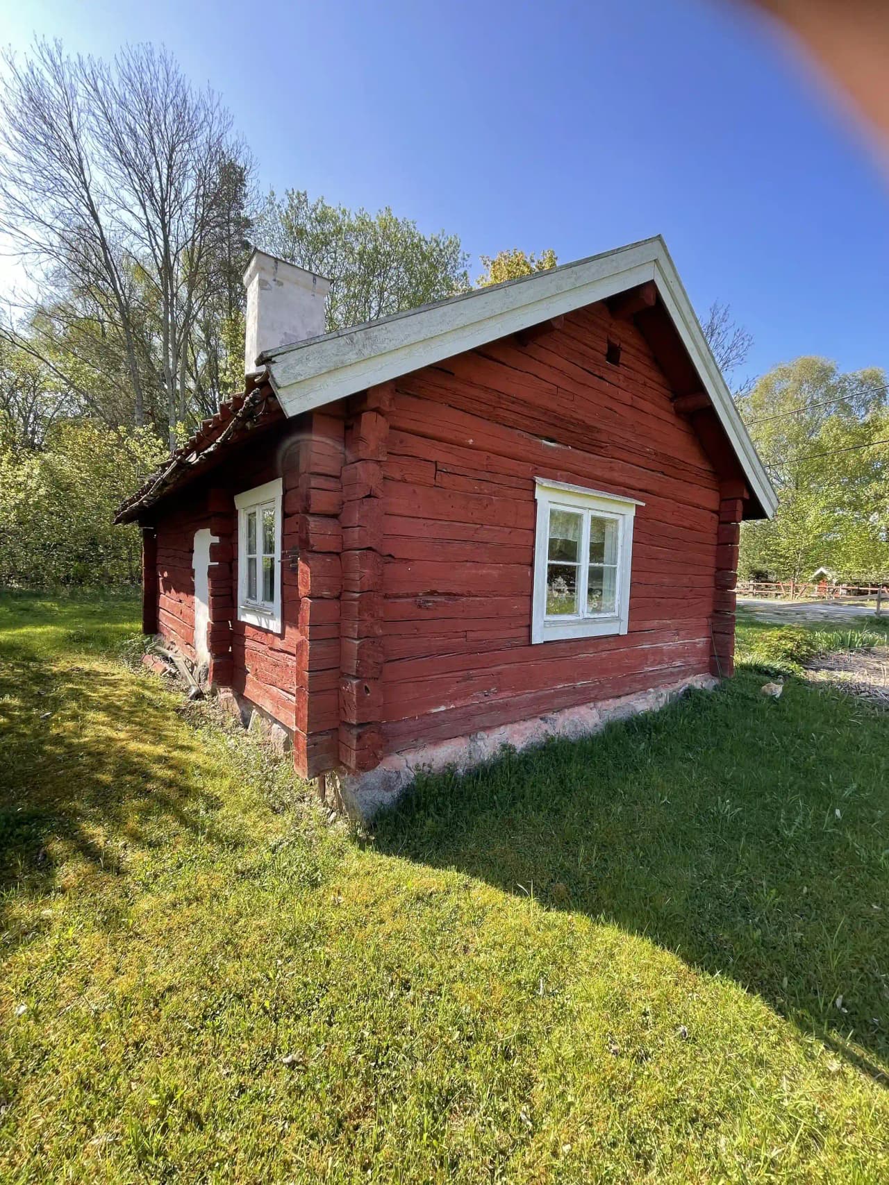 En traditionell hembygdsgård omgiven av grön gräsmatta, byggd i trä med tegeltak. Det ser ut som en lantlig stuga med en synlig dörr och del av en gårdsmiljö.