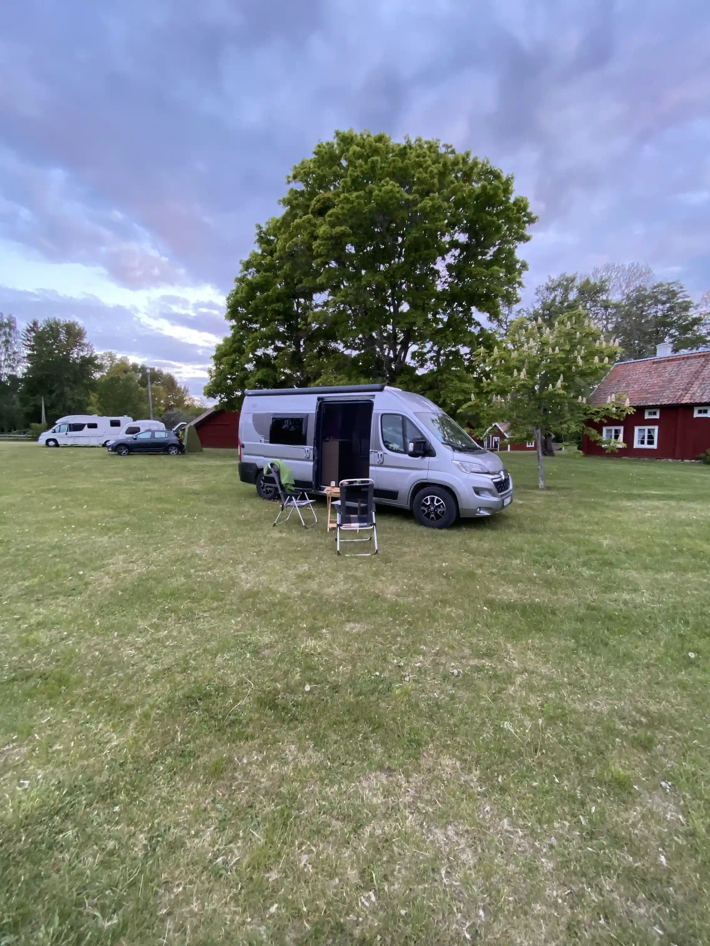 En husbil parkerad på en gräsmatta framför Kvarngården Hembygdsgård, omgiven av grönska.