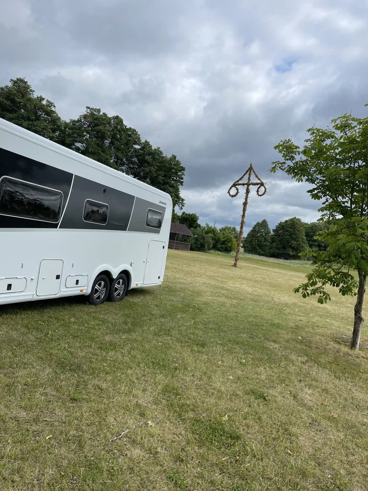 En bild av en hembygdsgård där flera olika typer av fordon, inklusive husvagn och hästtransport, är parkerade på grusplanen framför byggnaden.